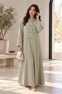 Model wearing The Jawahir Knotted Button Detail Closed Abaya in sage green, styled with a structured handbag for an elegant everyday look.