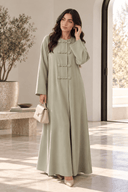 Model wearing The Jawahir Knotted Button Detail Closed Abaya in sage green, styled with a structured handbag for an elegant everyday look.
