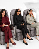 Three women seated in a row of gray airplane cabin seats, dressed in coordinated outfits, with one taking a notebook outtake and two resting with headphones on. The scene is set inside a bright airplane cabin.