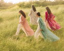 Four women walking through a sunlit grassy field, each wearing a flowing traditional outfit in soft pastel and bright colors including pink, green, red, and white.