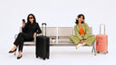 Two women sitting on a metal bench with their luggage in a clean studio setting. One woman wears a black outfit and sits next to a black hard-shell suitcase, while the other wears a green and orange outfit and sits next to a pink hard-shell suitcase.