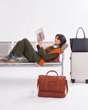 A woman reclines on a metal airport-style bench reading a magazine, with multiple travel bags around her: a dark tote bag on a light suitcase, and a brown leather briefcase-style bag in front. She wears an olive green outfit with an orange vest and blue socks.