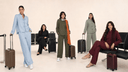 Five women modeling coordinated travel-themed outfits in light blue, black, olive green, sage green, and burgundy, each paired with a suitcase, posed in a minimalist beige studio setting with lounge chairs.