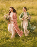 Two women running through tall grass holding flower bouquets, wearing coral striped and green geometric abayas over cream sets